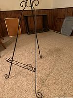 Full view of black metal easel standing on carpeted floor against wood-paneled wall