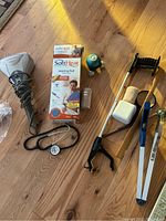 Photo showing handheld massager, boxed SoftHeat heating pad, stethoscope, grabber tool, blue clamp, small decorative item, and small plastic container on a wooden floor.