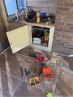 Overview of various hand tools and power tools including a red power drill, pliers, hammer, drill bit sets, wrench, and flexible vent hose scattered on tiled floor near an open cabinet.