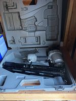 Open gray plastic carrying case with electric nail gun inside showing motor, handle, and nail magazine.