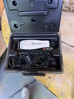 Open black plastic case showing Oster hair clipper, power cord, plastic guard combs, and cleaning brush inside.