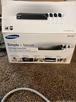 Box of Samsung Simple & Secure 4CH real-time DVR system showing DVR unit and four cameras, product details and branding.