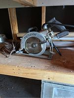 Photo shows a single Skilsaw circular saw placed on a wooden shelf. The saw is metallic with a black handle, a rotating saw blade partially covered by a metal guard with the brand name 'SKILSAW' written on it. Power cord is attached and coiled to the side.