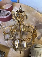 Ornate vintage metal hanging light fixture shown resting on a surface, displaying detailed floral and scroll metalwork and three candle-style white bulbs.