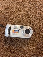 Silver Sony Cyber-shot digital camera lying on carpet, showing front with lens and logo, model shows 3.2 megapixels and 3x optical zoom details