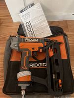 RIDGID straight finish nailer with attached nail magazine, placed on RIDGID branded soft carrying case along with instruction manual.