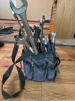 Blue fabric tool bag holding multiple various hand tools including wrenches, screwdrivers, pliers, and a hammer, placed on a wood floor.