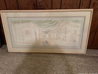 Framed wall art with a village scene, showing several buildings and a person in the center, set against a textured background, inside a light wooden frame and mat borders.