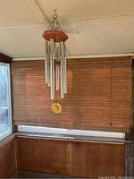 Wind chime hanging indoors against bamboo blinds and wooden paneling, showing complete structure including wooden top mount and six metal tubes.