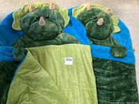 Two identical large dinosaur-themed slumber bags laid flat, showing the front and hood with dinosaur facial features and a light green inner lining.