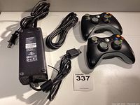 Two black wireless Xbox controllers, an AV cable adapter with Xbox proprietary connector, and a power adapter shown on a white surface.