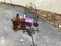 Photo of assorted home decor items including two brown decorative vases, one glass vase, one tall white ceramic vase, a wrapped package of two purple bath towels, a metal stand, and metal wall art pieces on asphalt surface.