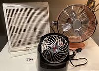 Three small electric fans on a white surface: a Holmes white box fan, a black Vornado desk fan, and a copper-colored metal blade fan.