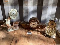 Photo displaying three clocks together on a wooden surface under sunlight; from left the resin angel clock, center boat-shaped Sessions windup clock, right the glass dome clock.