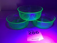 Three round uranium glass bowls glowing green under UV light, stacked in triangular formation with no visible chips or cracks.