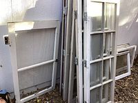 Multiple wood-framed windows leaning against exterior wall, including multi-pane and single-pane styles