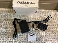 Personal air purifier device with strap and power adapter laid out on carpet with original box in background