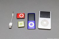 All four Apple iPods and the USB to headphone adapter laid on a gray background, showing front views including screens and click wheels/buttons.