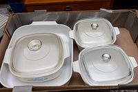Four CorningWare dishes including one large Shadow Iris rectangular pan and three smaller Blue Cornflower casserole dishes with lids inside a cardboard box.
