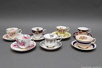 Front view of seven assorted bone china teacups and saucers showing various floral and decorated patterns with gold accents.