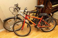 Both mountain bikes shown side by side indoors on a wooden floor against a beige wall, highlighting their frames and tires.