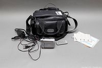 Full lot including Canon Elura-100 camcorder, power cord, remote control, instruction manuals and carrying case displayed on grey surface.
