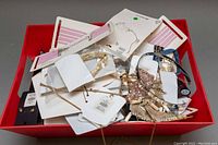 Red plastic bin containing assorted new costume jewelry items such as necklaces, chokers, earrings with some on display cards, showing mixed materials like shells, beads, sequins, and metal chains.