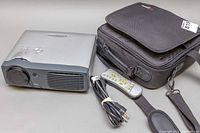 Front angled view of silver-gray Optoma EP739 projector, remote, power cable, and black carrying case with shoulder strap.