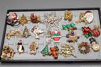 Full tray view showing assortment of over 20 vintage Christmas-themed brooches including Santa, snowflakes, trees, animals, and festive icons.