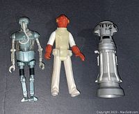 Three Star Wars action figures laid side by side. Figures include a silver humanoid robot, an orange alien in beige and white clothes, and a tall silver-gray droid.