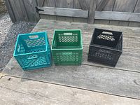 Three plastic milk crates in teal, green, and black colors placed on a wooden surface outside, showing their overall design and condition.