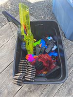 Black plastic bin containing various toy blasters and accessories including a translucent yellow dart gun, a red and black toy gun, and a plastic belt of toy ammunition rounds.