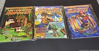 Three Nintendo Power magazines laid flat, covers visible, showing Donkey Kong 64, Mario Party 2 with Mario and Yoshi, and a classic Mario cover with text about tips and cheats.