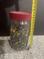 Full jar of vintage cats eye marbles with red lid, seen next to measuring tape showing about 15 cm height