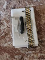 Photo of a metal Apple Watch bracelet in silver and gold tone, placed on a white cardboard backing with a sealed small black tool in plastic.