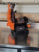 Photo of a medium-sized bench-top belt sander with orange and black motor housing on a metal surface, showing power cord, sanding belt, worktable, and switch.