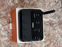 Black Bluetooth transmitter & receiver device with three buttons and antenna, placed on top of its box showing mode indicators.
