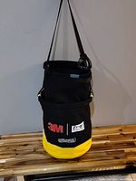 Photo of black and yellow canvas bucket with attached strap and safe hook, showing logos of 3M, SALA, and DeWalt.