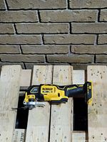 Dewalt cordless multitool laying on wooden pallets against brick wall showing tool body and attachment area. Yellow and black body with 20V XR branding visible.