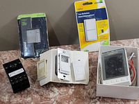 Shows various electrical control devices including a programmable dimmer with remote, a thermostat, a circuit breaker, and two wall switch units in packaging.