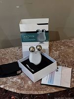 Facial toning device displayed in open box with included charging cable, carrying pouch, cleaning cloth, and instruction manual on marble surface.