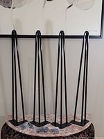 Four black metal hairpin table legs, 28 inches tall, with mounting plates and hardware on a patterned table surface, positioned against a wall with artwork.