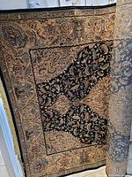 Partially rolled carpet showing the inner and outer patterns, black central field with intricate floral designs, bordered by tan and rust colored detailed patterns.