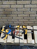 Four bar clamps on wood pallets against brick wall background with two yellow and black DeWalt clamps, one blue and black Firepower clamp, and one red clamp.