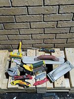 Photo showing a variety of hand tools arranged on a wooden surface against a brick wall including trowels, utility knives, scrapers, and a paint roller handle.