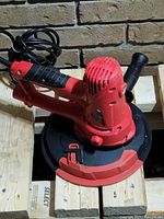 Photo shows a red and black handheld electric orbital sander with attached power cord and side handle resting on a wooden surface.