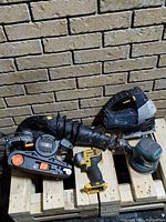 Five power tools arranged on wooden pallets against a brick wall: belt sander, cordless drill, jigsaw, detail sander, orbital sander