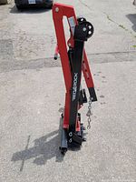 Folded red and black hydraulic engine hoist standing on pavement; boom with hook and chain, hydraulic cylinder and caster wheels visible
