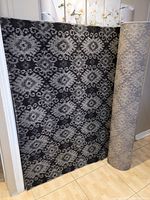 Photo showing black and grey Tuscany rug with intricate symmetrical floral and geometric motifs, partially rolled up against a wall on tile flooring.