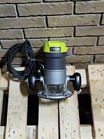 Ryobi router tool placed on a wooden pallet, showing front view with power cord coiled beside it.